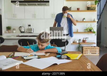 Caucasian man at home with his son together, in kitchen, boy doing homework at table. Social distancing during Covid 19 Coronavirus quarantine lockdow Stock Photo
