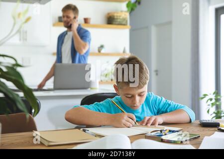 Caucasian man at home with his son together, in kitchen, boy doing homework at table. Social distancing during Covid 19 Coronavirus quarantine lockdow Stock Photo