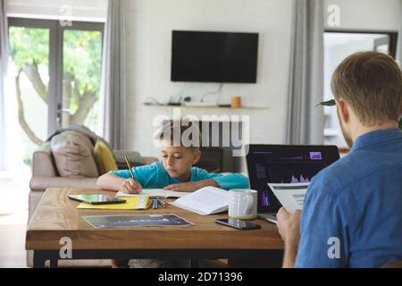 Caucasian man at home with his son together, at table in kitchen, father working using laptop computer, boy doing homework. Social distancing during C Stock Photo