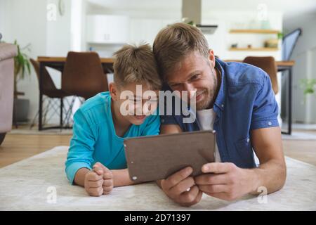 Caucasian man at home with his son together, lying on rug in living room, using digital tablet, smiling. Social distancing during Covid 19 Coronavirus Stock Photo