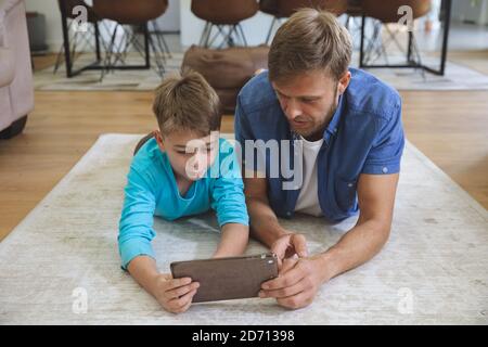 Caucasian man at home with his son together, lying on rug in living room, using digital tablet. Social distancing during Covid 19 Coronavirus quaranti Stock Photo