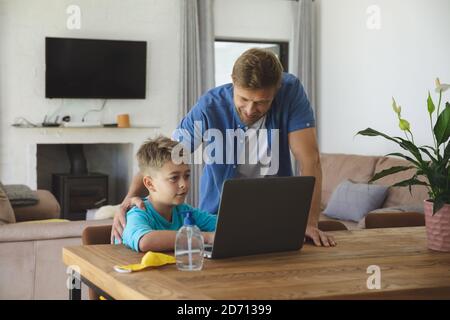 Caucasian man at home with his son together, boy sitting at table, using laptop computer, father standing next to him. Social distancing during Covid Stock Photo
