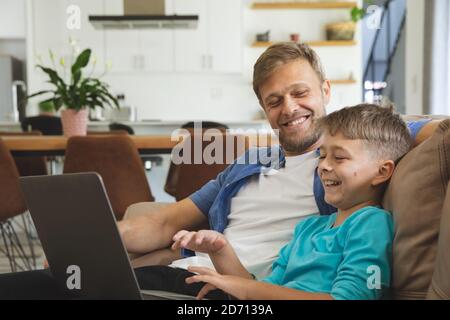 Caucasian man at home with his son together, sitting on sofa in living room, using laptop computer, smiling. Social distancing during Covid 19 Coronav Stock Photo