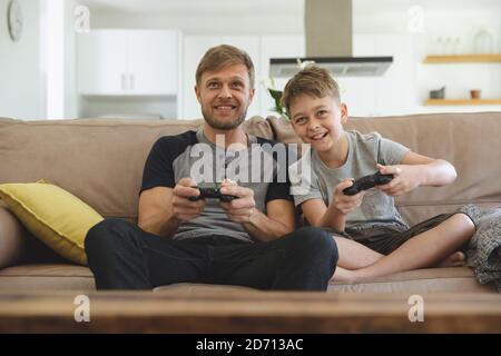 Caucasian man at home with his son together, sitting on sofa in living room, playing video games, smiling. Social distancing during Covid 19 Coronavir Stock Photo