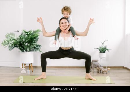 Mother and son doing fitness yoga exercise at home. Charming family spends time in the gym, mother stands in yoga pose and little cute boy kid sitting Stock Photo