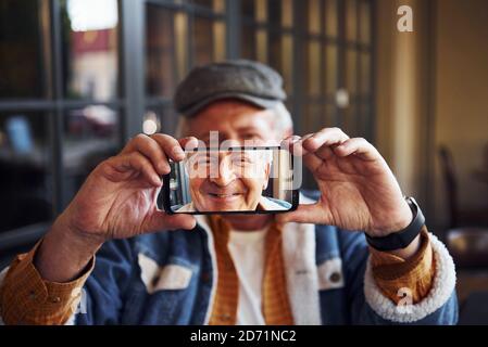 Stylish senior in fashionable clothes and in glasses sits in the cafe and holds phone against his face Stock Photo