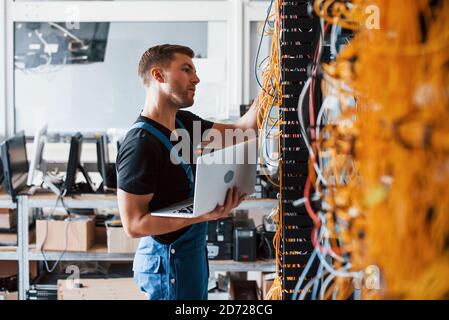 Young man in uniform and with laptop works with internet equipment and wires in server room Stock Photo