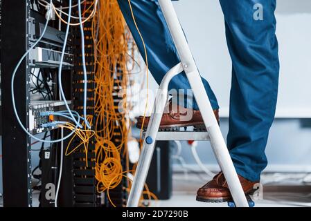 Man in uniform stands on the ladder and works with internet equipment and wires in server room Stock Photo