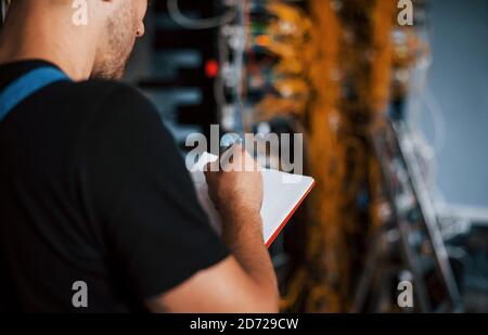 Young man in uniform with notepad in hands works with internet equipment and wires in server room Stock Photo