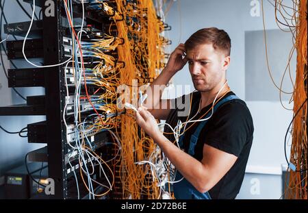 Young man in uniform feels confused and looking for a solution with internet equipment and wires in server room Stock Photo
