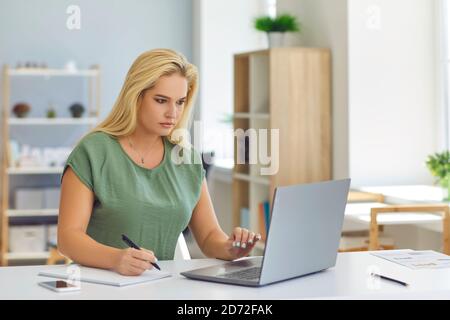 Woman sitting at desk, working online with laptop and making notes Stock Photo