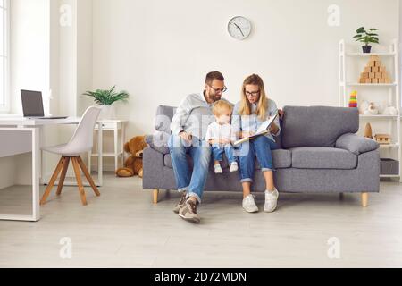 Mom shows pictures in a book to her little son, who is sitting with her and dad on the couch in the room. Stock Photo