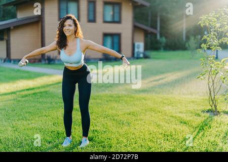 Slim motivated brunette woman dressed in cropped top and leggings, has workout with dumbbells, poses on green lawn near private house, has perfect bod Stock Photo