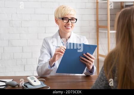 Friendly smiling adult female doctor in glasses, white coat makes notes on tablet and looks at lady Stock Photo