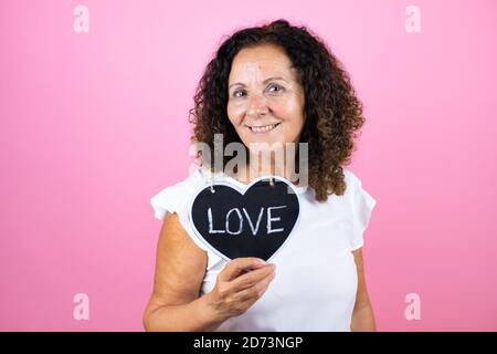 Middle age woman wearing casual white shirt standing over isolated pink background smiling and holding heart blackboard with love word message Stock Photo