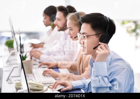 Asian helpdesk representative in headphones with his colleagues providing tech support services to customers, copy space Stock Photo