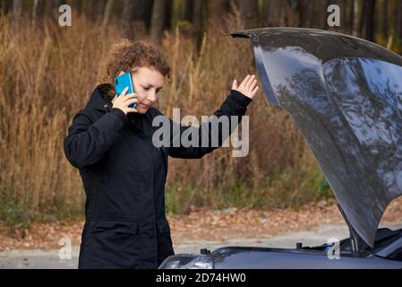 An upset woman stands near her broken car and talking on a cell phone on the road in autumn. Stock Photo