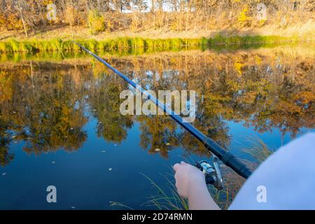 Hand of woman fisherman with fishing rod and spinning reel on river bank. Concept of fishing as sport and hobby. Selective focus, autumn landscape. Stock Photo