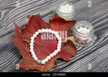 A beaded bracelet on a dried autumn maple leaf. Nearby are jars of beads. On pine planks in black. Stock Photo