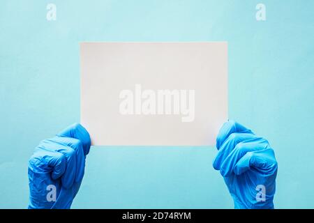 Hands in blue latex gloves holding blank paper card on on blue background. Medical poster template Stock Photo