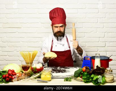 Chef makes dough. Italian cuisine concept. Man with beard holds dough and rolling pin on white brick background. Cook with strict face in burgundy uniform sits by table with vegetables and kitchenware Stock Photo
