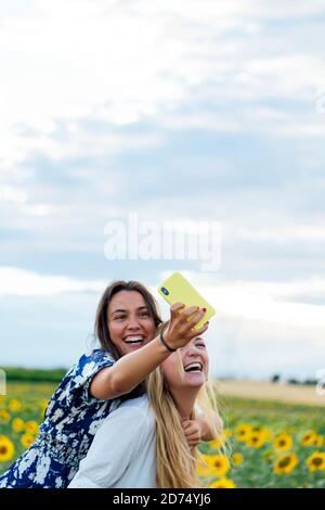 A couple of attractive young women one blonde and the other brunette posing in their designer dresses in a field of sunflowers using their smartphone Stock Photo
