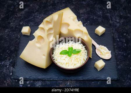 Two wedge-shaped pieces of dutch cheese and a bowl of grated cheese with basil leaves on a black slate stone board