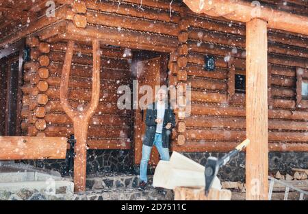 Man staying in the country house porch and enjoying snowfall. Wooden stump with axe on snowy yard unfocused with heavy snowflakes . Winter countryside Stock Photo