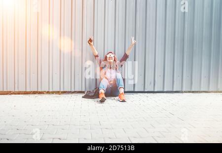 Cheerful hands rising UP Beautiful modern smiling young female teenager in a checkered shirt and jeans with headphones and smartphone Stock Photo