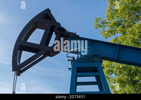 Industrial block pumping unit with blue framed body and black rocking element with flexible rope against cloudy blue sky and green tree foliage Stock Photo