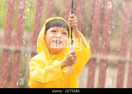 Happy Asian little child holding an umbrella having fun playing with the raindrops. A boy wearing a yellow raincoat. Stock Photo