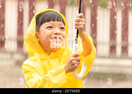 Happy Asian little child holding an umbrella having fun playing with the raindrops. A boy wearing a yellow raincoat. Stock Photo