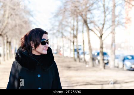 Young woman in a black coat, dark sunglasses and earrings walks along a city street, illuminated by bright spring sunlight. Stock Photo
