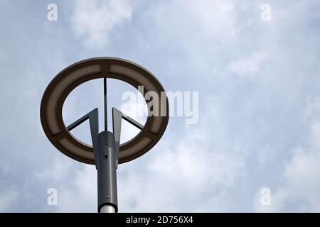A grey round lamp with an led base with black three branched holders mounted on a cylindrical lamp post against a blue sky covered with white clouds Stock Photo