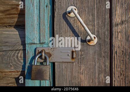Old metal rusty lock on wooden door with white-painted, battered handle bolted on screws locked through bracket to door frame on bolt of log structure Stock Photo