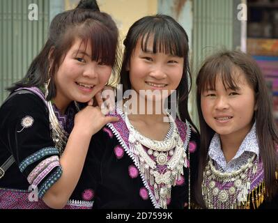 Three Vietnamese Hmong teenage girls wear their clan’s traditional embroidered Sunday Best with heavy silver necklaces and smile for the camera. Stock Photo