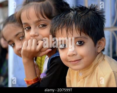 Three little Indian Rajasthani children with traditional kohl eyeliner sit in a row and pose for the camera. Stock Photo