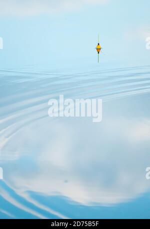 A calm surface of water reflecting a blue sky with cumulus clouds crossing several circles of small waves and a defocused yellow fishing float in the Stock Photo