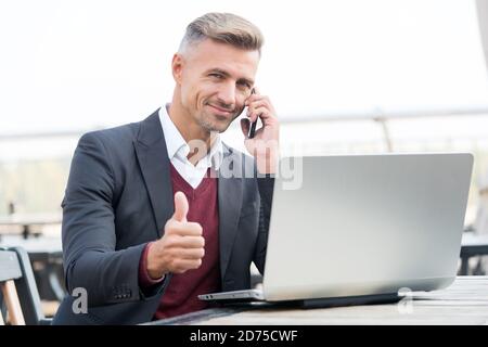 Happy businessman show thumbs up hand gesture talking to client on mobile phone and using computer for business online, approval. Stock Photo