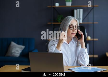 Smiling senior grey-haired beautiful businesswoman talking on the phone in office. Business portrait of attractive mature lady wearing eyeglasses Stock Photo