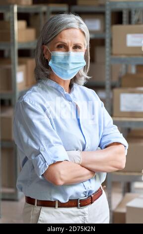 Older woman entrepreneur wearing face mask standing in warehouse, portrait. Stock Photo