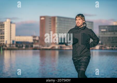 Winter run man jogging outdoor in city harbour running outside wearing cold weather accessories - hat ,gloves , windproof sport jacket. Active fit Stock Photo