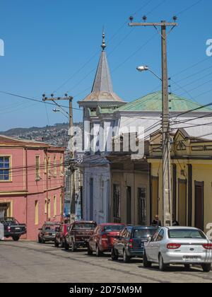 Valparaiso, Chile - December 8, 2008: Lower class neighborhood street with a few houses, one with tower, and cars parked in front under blue sky. Stock Photo