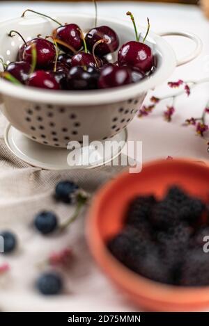 Cherries in a strainer and some blackberries on a bowl out of focus on a pink background and a cloth Stock Photo