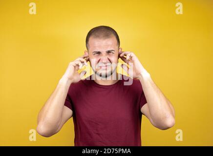 Russian man wearing basic red t-shirt over yellow insolated background covering ears with fingers with annoyed expression for the noise of loud music. Stock Photo