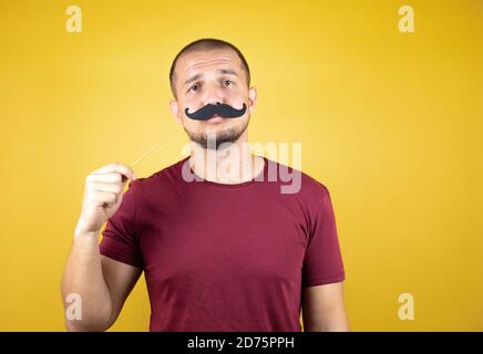 Russian man wearing basic red t-shirt over yellow insolated background happy with mustache mask Stock Photo