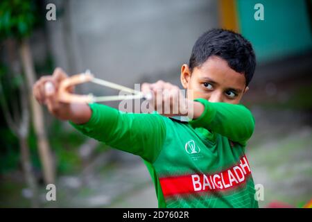 Bangladesh – October 04, 2020: A Bangladesh cricket jersey-wearing kid boy shooting a wooden slingshot at Barinagar, Jessore. Stock Photo