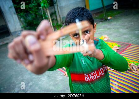 Bangladesh – October 04, 2020: A Bangladesh cricket jersey-wearing kid boy shooting a wooden slingshot at Barinagar, Jessore. Stock Photo