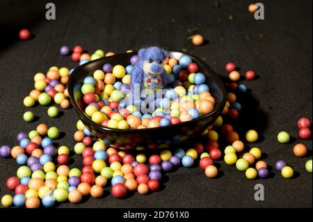 Cute blue toy bear inside a black bowl filled with colorful candies over black background Stock Photo