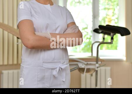 Closeup of a doctor in a white uniform, with folded hands in front of him. Stands in front of a gynecological chair. Stock Photo